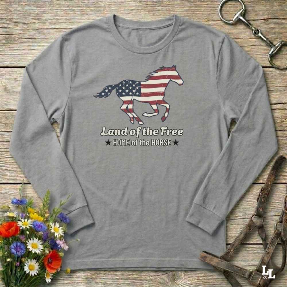 Gray long-sleeve shirt with American flag horse design on a wooden surface with flowers and equestrian equipment.