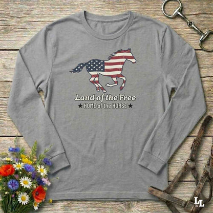 Gray long-sleeve shirt with American flag horse design on a wooden surface with flowers and equestrian equipment.