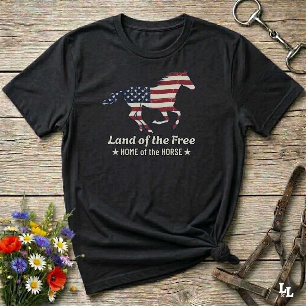 black t-shirt lying on wooden floor, graphic horse design on t-shirt of running horse with american flag, and the words Land of the Free, Home of the Horse. Surrounded by horse objects and some flowers.