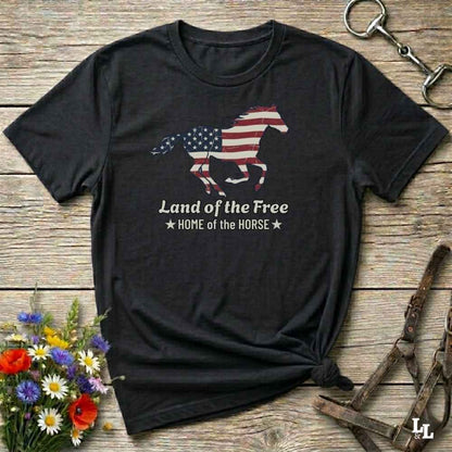 black t-shirt lying on wooden floor, graphic horse design on t-shirt of running horse with american flag, and the words Land of the Free, Home of the Horse. Surrounded by horse objects and some flowers.