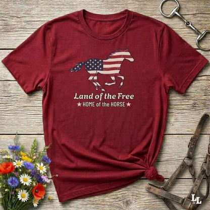 red t-shirt lying on wooden floor, graphic horse design on t-shirt of running horse with american flag, and the words Land of the Free, Home of the Horse. Surrounded by horse objects and some flowers.