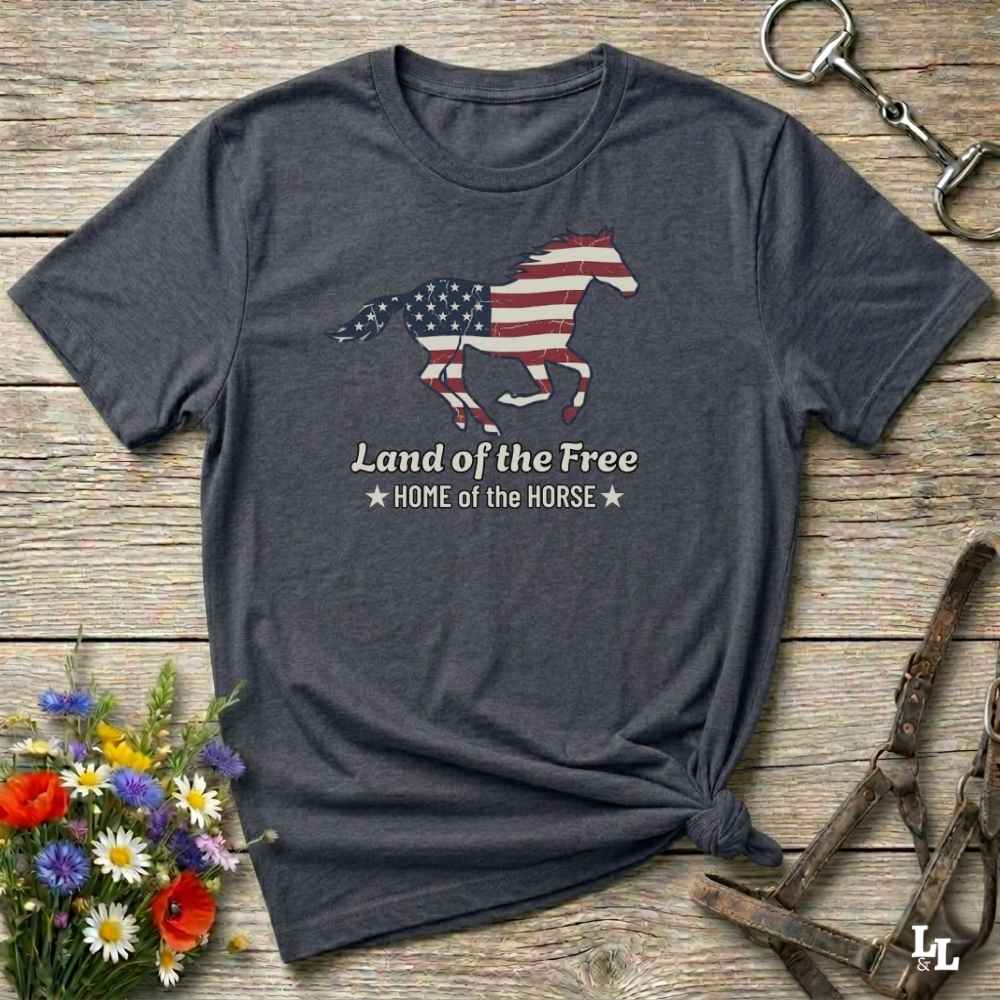 gray t-shirt lying on wooden floor, graphic horse design on t-shirt of running horse with american flag, and the words Land of the Free, Home of the Horse. Surrounded by horse objects and some flowers.