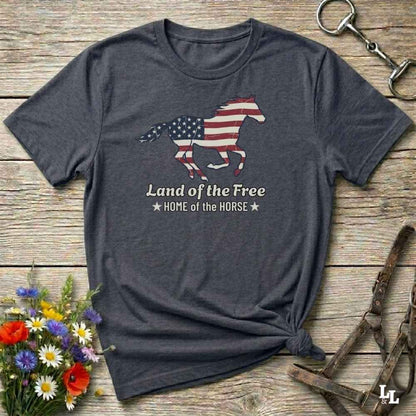 gray t-shirt lying on wooden floor, graphic horse design on t-shirt of running horse with american flag, and the words Land of the Free, Home of the Horse. Surrounded by horse objects and some flowers.