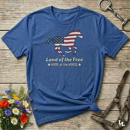 blue t-shirt lying on wooden floor, graphic horse design on t-shirt of running horse with american flag, and the words Land of the Free, Home of the Horse. Surrounded by horse objects and some flowers.