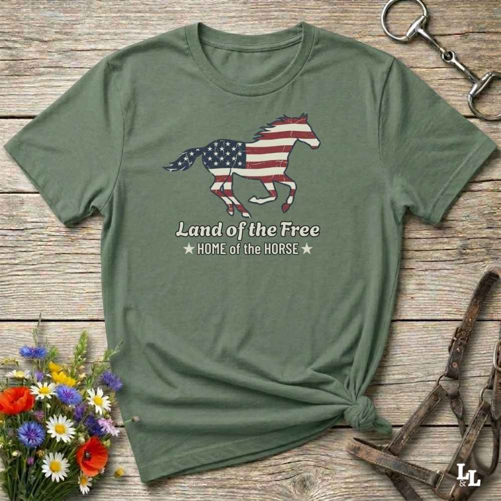 green t-shirt lying on wooden floor, graphic horse design on t-shirt of running horse with american flag, and the words Land of the Free, Home of the Horse. Surrounded by horse objects and some flowers.