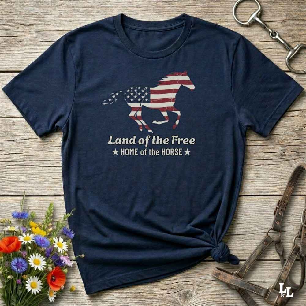 navy blue t-shirt lying on wooden floor, graphic horse design on t-shirt of running horse with american flag, and the words Land of the Free, Home of the Horse. Surrounded by horse objects and some flowers.