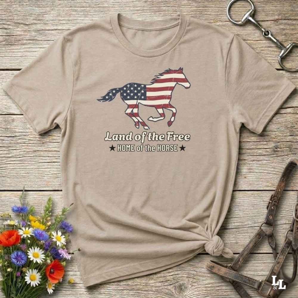 beige t-shirt lying on wooden floor, graphic horse design on t-shirt of running horse with american flag, and the words Land of the Free, Home of the Horse. Surrounded by horse objects and some flowers.