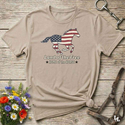 beige t-shirt lying on wooden floor, graphic horse design on t-shirt of running horse with american flag, and the words Land of the Free, Home of the Horse. Surrounded by horse objects and some flowers.
