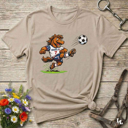 Beige t-shirt with a cartoon horse kicking a soccer ball on a wooden surface with flowers and horse halter and bit.