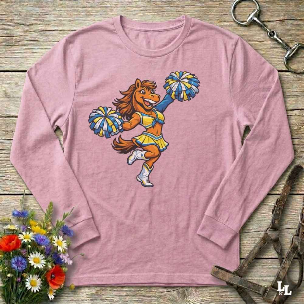 Pink long-sleeve shirt with a cartoon horse cheerleader design on a wooden surface with flowers and a bridle.