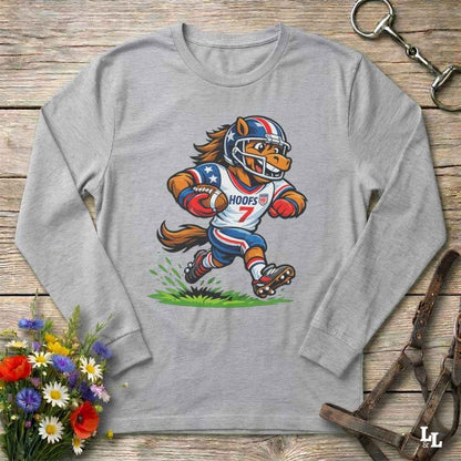 A light gray long-sleeve t-shirt with a cartoon graphic of a horse mascot in a football uniform, styled on a rustic wooden background with flowers and leather tack.