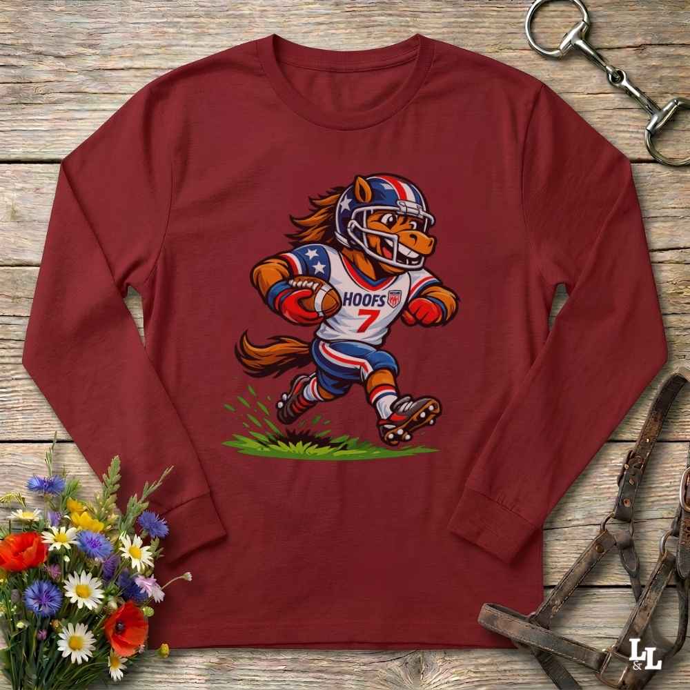 A long-sleeve maroon crewneck shirt laid flat on a rustic wood surface, featuring a cartoon horse mascot in a football uniform with the word 'HOOFS' on the front. The shirt is styled with flowers, a metal tack item, and leather straps around it.