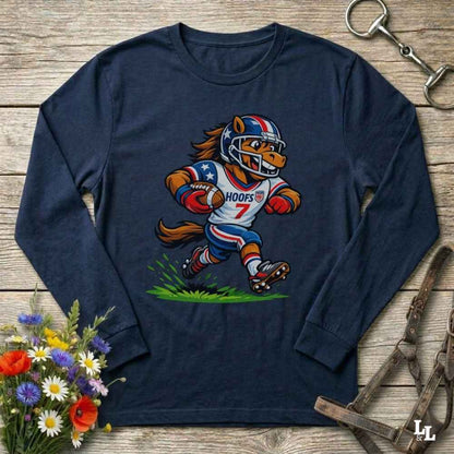 A navy blue long-sleeve t-shirt with a cartoon graphic of a horse running as a football player wearing a red, white, and blue helmet. The shirt is displayed flat on a rustic wooden background with decorative flowers and horse tack.