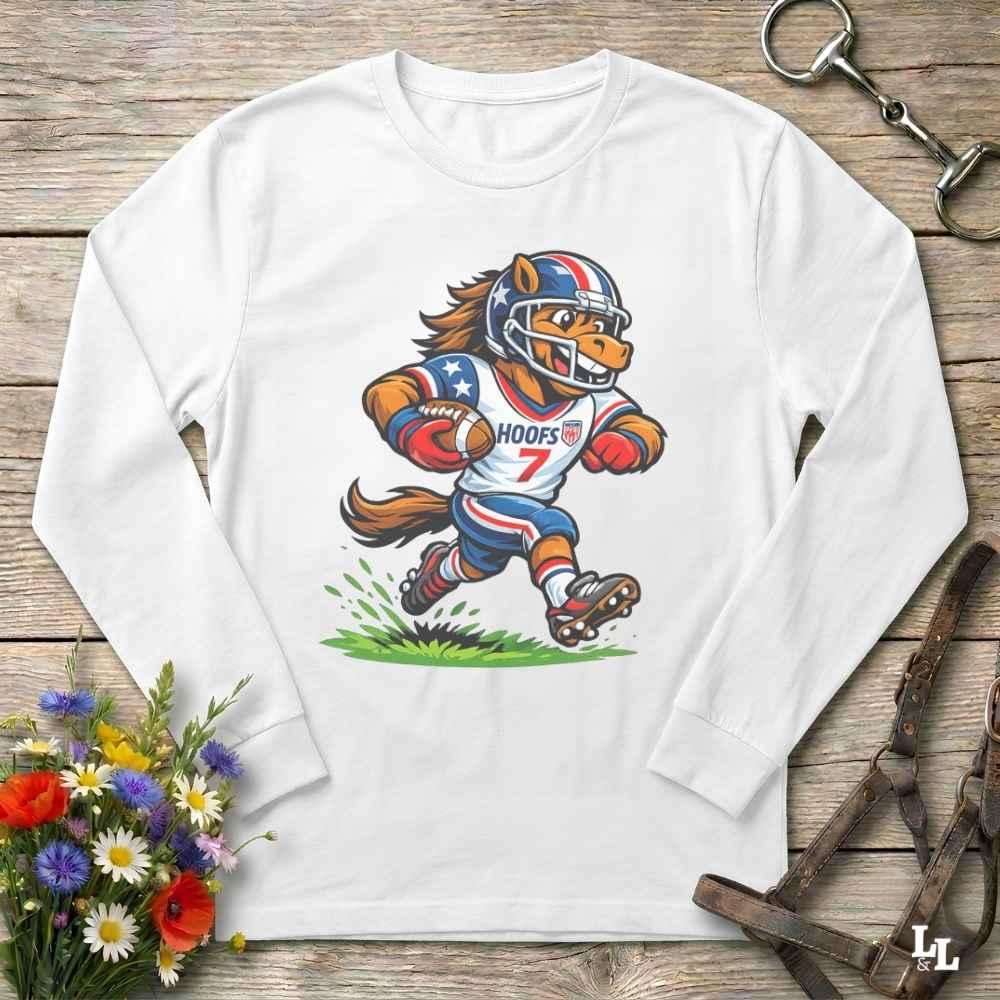 A white long-sleeve crewneck t-shirt laid flat on a rustic wooden surface, featuring a colorful cartoon graphic of a horse mascot in a football uniform. The shirt is styled with equestrian accessories (a leather bridle and a horse bit) and a small bouquet of colorful wildflowers.