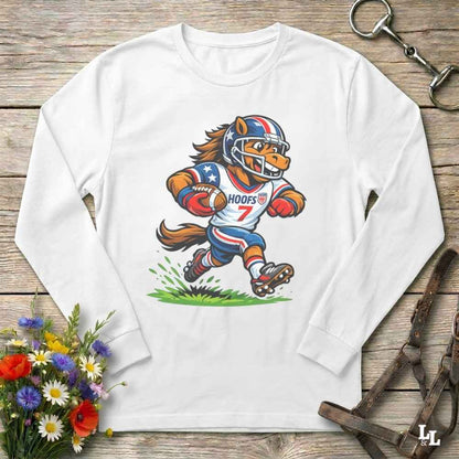 A white long-sleeve crewneck t-shirt laid flat on a rustic wooden surface, featuring a colorful cartoon graphic of a horse mascot in a football uniform. The shirt is styled with equestrian accessories (a leather bridle and a horse bit) and a small bouquet of colorful wildflowers.