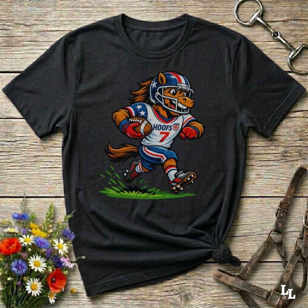 A black short-sleeve t-shirt with a cartoon graphic of a horse football player wearing a red, white, and blue helmet and jersey with the name 'HOOFS' and number '7'. The shirt is styled flat on a rustic wooden surface with decorative flowers and leather 