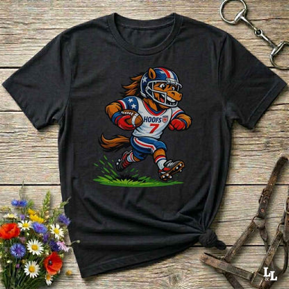 A black short-sleeve t-shirt with a cartoon graphic of a horse football player wearing a red, white, and blue helmet and jersey with the name 'HOOFS' and number '7'. The shirt is styled flat on a rustic wooden surface with decorative flowers and leather 