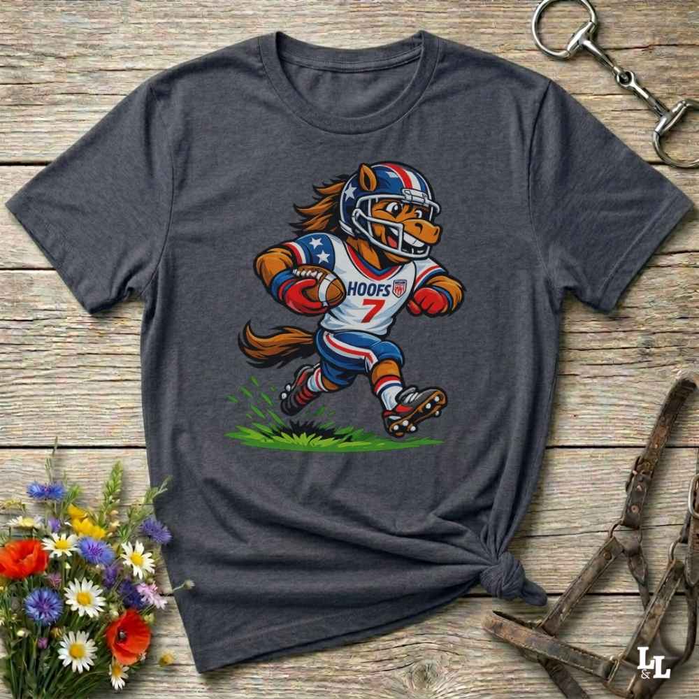 A dark heather grey short-sleeve t-shirt with a cartoon graphic of a horse football player wearing a red, white, and blue helmet and jersey with the name 'HOOFS' and number '7'. The shirt is styled flat on a rustic wooden surface with decorative flowers and leather 