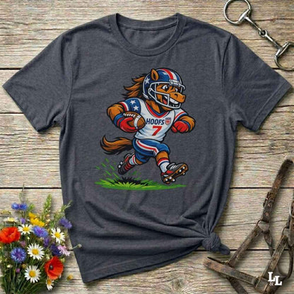 A dark heather grey short-sleeve t-shirt with a cartoon graphic of a horse football player wearing a red, white, and blue helmet and jersey with the name 'HOOFS' and number '7'. The shirt is styled flat on a rustic wooden surface with decorative flowers and leather 
