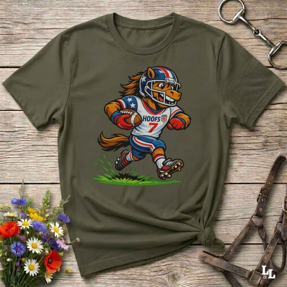 A green short-sleeve t-shirt with a cartoon graphic of a horse football player wearing a red, white, and blue helmet and jersey with the name 'HOOFS' and number '7'. The shirt is styled flat on a rustic wooden surface with decorative flowers and leather 