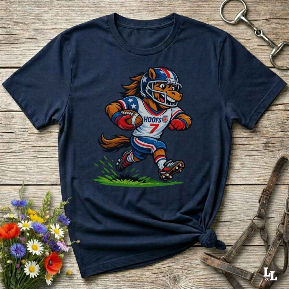 A navy blue short-sleeve t-shirt with a cartoon graphic of a horse football player wearing a red, white, and blue helmet and jersey with the name 'HOOFS' and number '7'. The shirt is styled flat on a rustic wooden surface with decorative flowers and leather straps.