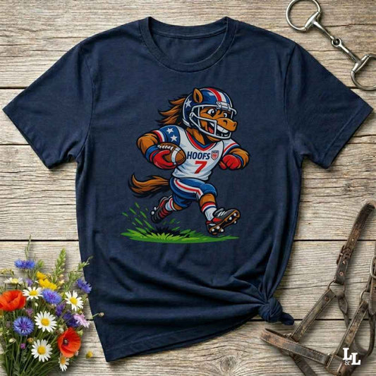 A navy blue short-sleeve t-shirt with a cartoon graphic of a horse football player wearing a red, white, and blue helmet and jersey with the name 'HOOFS' and number '7'. The shirt is styled flat on a rustic wooden surface with decorative flowers and leather straps.