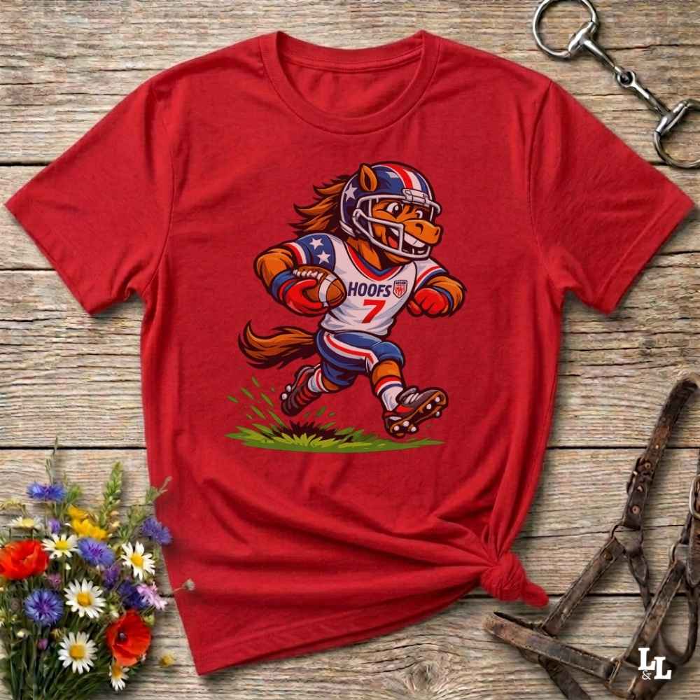 A red short-sleeve t-shirt with a cartoon graphic of a horse football player wearing a red, white, and blue helmet and jersey with the name 'HOOFS' and number '7'. The shirt is styled flat on a rustic wooden surface with decorative flowers and leather 