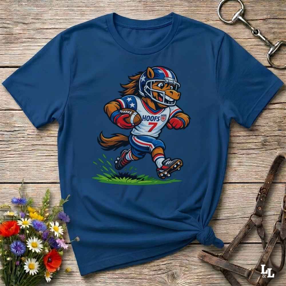 A blue short-sleeve t-shirt with a cartoon graphic of a horse football player wearing a red, white, and blue helmet and jersey with the name 'HOOFS' and number '7'. The shirt is styled flat on a rustic wooden surface with decorative flowers and leather 