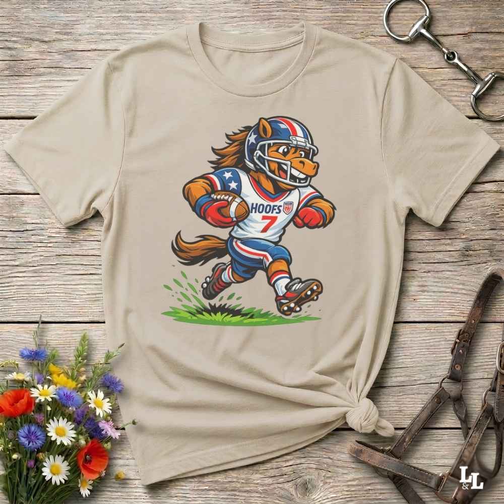 A beige short-sleeve t-shirt with a cartoon graphic of a horse football player wearing a red, white, and blue helmet and jersey with the name 'HOOFS' and number '7'. The shirt is styled flat on a rustic wooden surface with decorative flowers and leather 