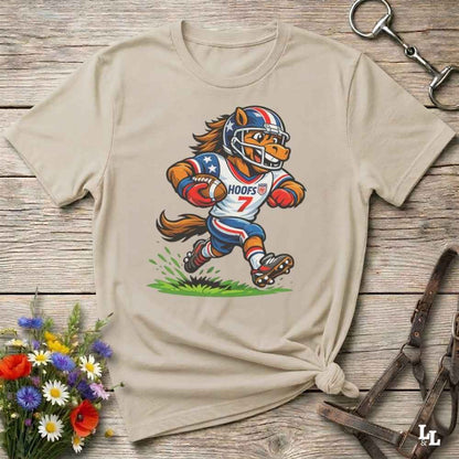 A beige short-sleeve t-shirt with a cartoon graphic of a horse football player wearing a red, white, and blue helmet and jersey with the name 'HOOFS' and number '7'. The shirt is styled flat on a rustic wooden surface with decorative flowers and leather 