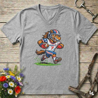 A heather gray V-neck t-shirt laid flat on a rustic wooden surface, featuring a cartoon graphic of a horse mascot in a football uniform with the number 7. The shirt is styled with equestrian tack in the background and a small bouquet of flowers in the lower-left corner.