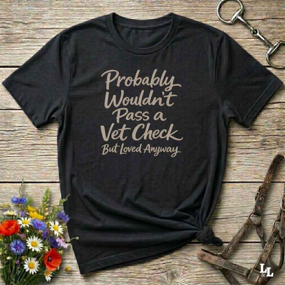 A black short-sleeve graphic T-shirt laid flat on a rustic wooden surface, with the phrase 'Probably Wouldn't Pass a Vet Check But Loved Anyways' printed in a light beige script. The shirt is styled with flowers, a horse bit, and a leather halter.