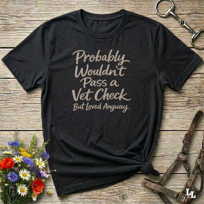 A black short-sleeve graphic T-shirt laid flat on a rustic wooden surface, with the phrase 'Probably Wouldn't Pass a Vet Check But Loved Anyways' printed in a light beige script. The shirt is styled with flowers, a horse bit, and a leather halter.