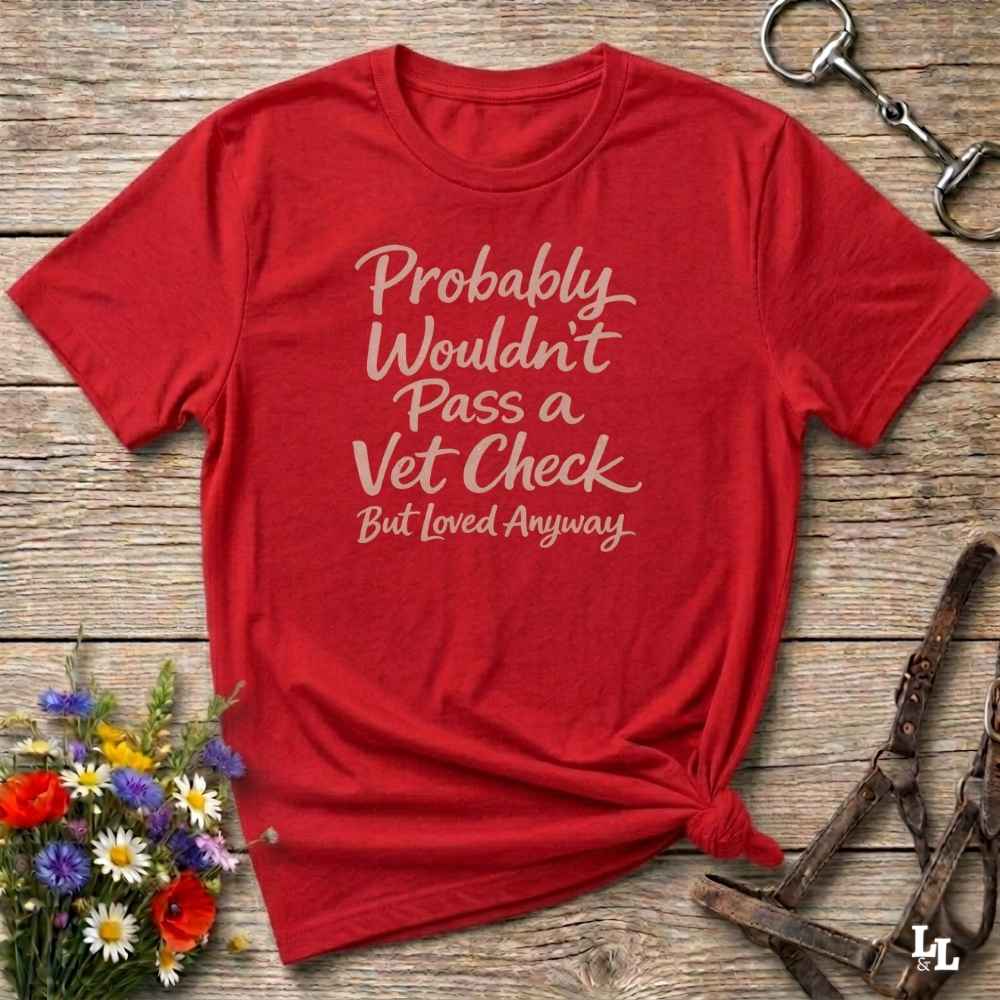 A red short-sleeve graphic T-shirt laid flat on a rustic wooden surface, with white script text reading 'Probably Wouldn't Pass a Vet Check But Loved Anyway.' Decorative flowers and equestrian-style accessories are placed around it as styling props.