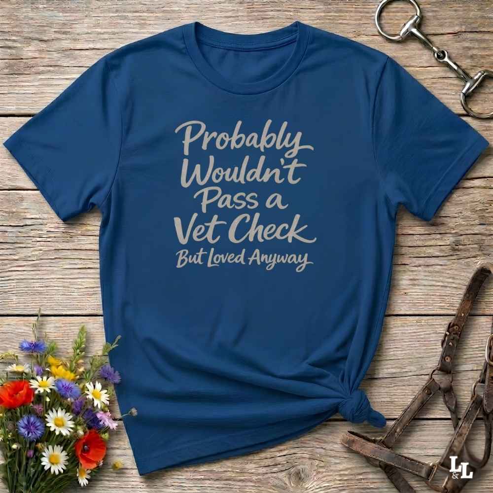 A navy blue graphic t-shirt with the text 'Probably Wouldn't Pass a Vet Check But Loved Anyways' in white script, styled in a flat-lay on a rustic wooden background with equestrian accessories and flowers.