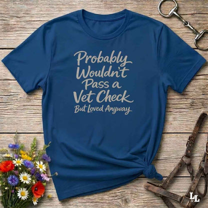 A navy blue graphic t-shirt with the text 'Probably Wouldn't Pass a Vet Check But Loved Anyways' in white script, styled in a flat-lay on a rustic wooden background with equestrian accessories and flowers.