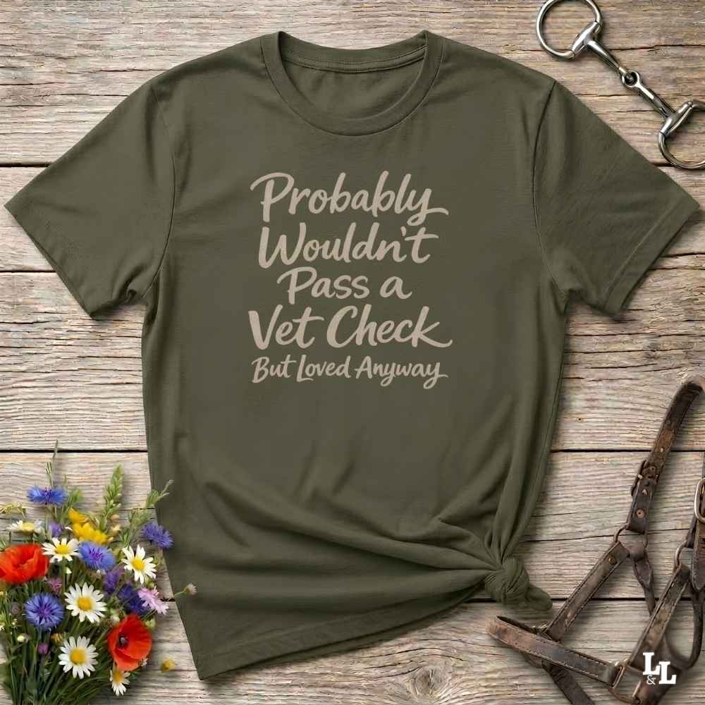 An olive green graphic t-shirt with the text 'Probably Wouldn't Pass a Vet Check But Loved Anyway' in a light beige script font. The t-shirt is styled in a flat-lay arrangement on a rustic wooden background, surrounded by equestrian-themed props including a horse bit, leather bridle straps, and a small bouquet of colorful wildflowers.