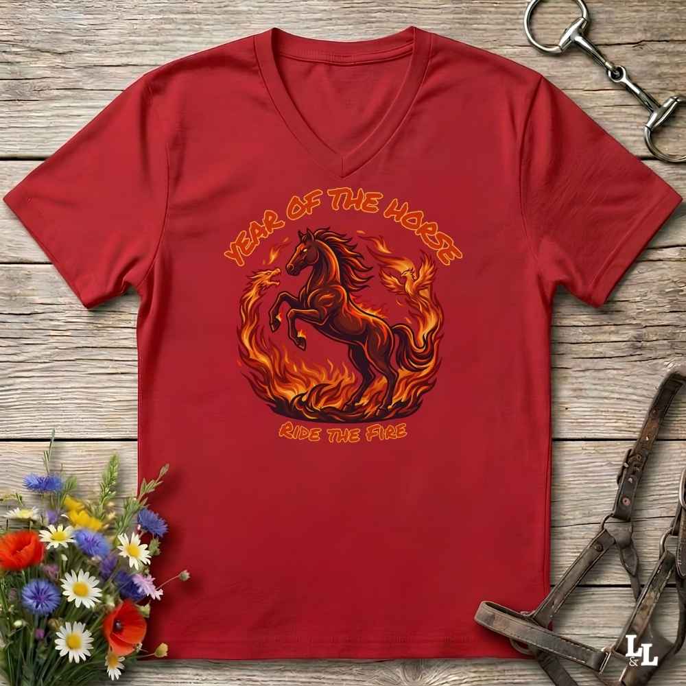 red V-neck t-shirt laid flat on a rustic wooden surface, featuring a graphic of a rearing horse surrounded by flames with the text 'YEAR OF THE HORSE' and 'RIDE THE FIRE'. The shirt is styled with equestrian props including a leather bridle, horse bits, and a small bouquet of wildflowers.