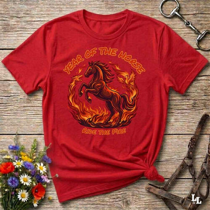 A red short-sleeve graphic T-shirt laid flat on a rustic wooden surface, featuring a fiery horse design with the text 'YEAR OF THE HARE' and 'RIDE THE FIRE.' Decorative flowers and equestrian hardware are placed around the shirt.