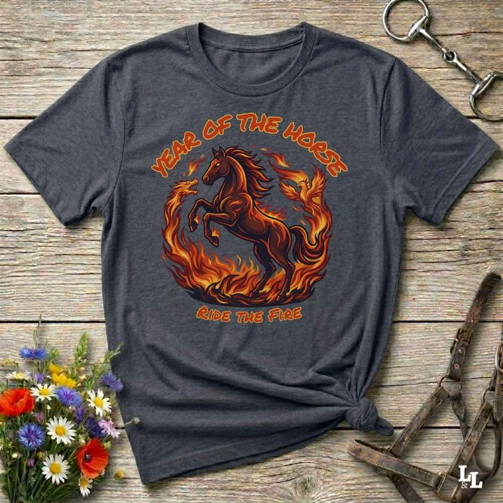 A dark heather gray graphic t-shirt featuring a fiery red and orange horse design with the text 'YEAR OF THE HORSE' and 'RIDE THE FIRE'. The shirt is styled in a flat-lay on a rustic wooden background with horse tack and flowers.