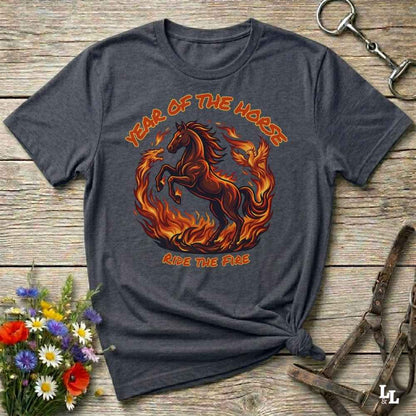 A dark heather gray graphic t-shirt featuring a fiery red and orange horse design with the text 'YEAR OF THE HORSE' and 'RIDE THE FIRE'. The shirt is styled in a flat-lay on a rustic wooden background with horse tack and flowers.