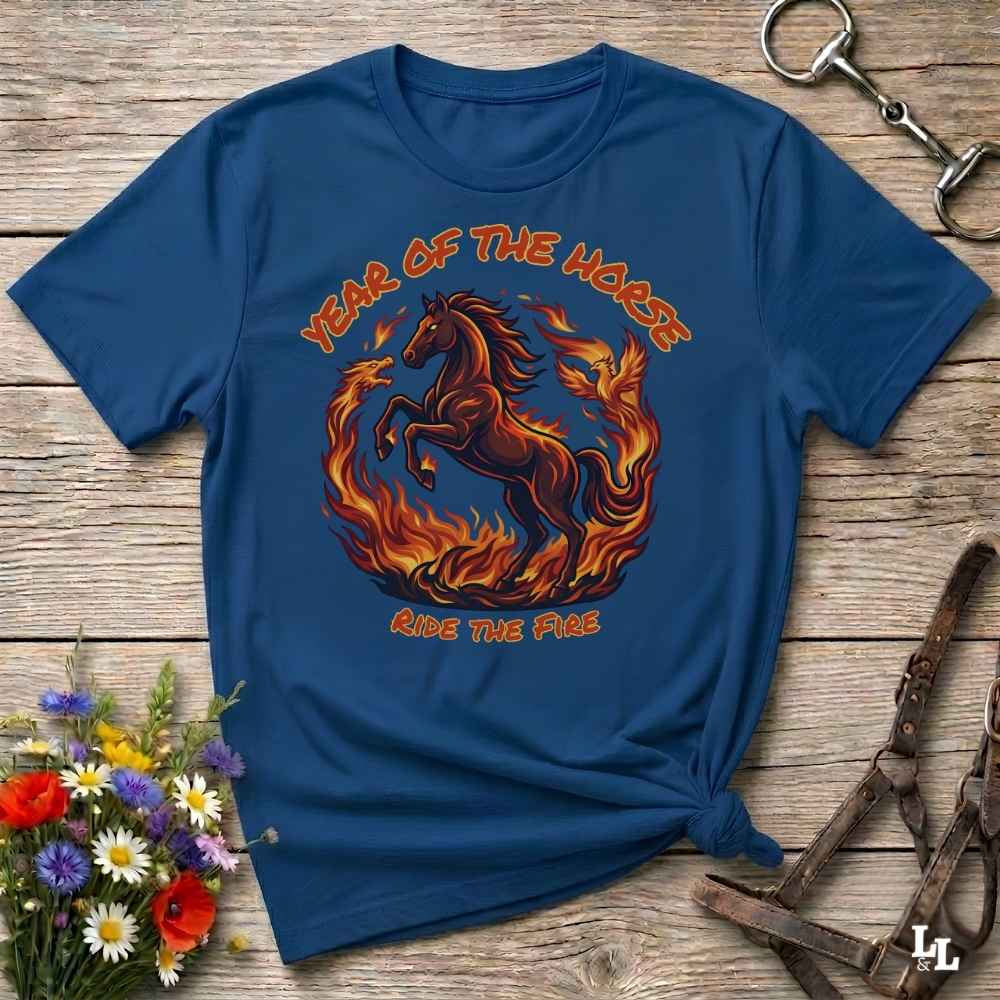 A navy blue graphic t-shirt featuring a fiery horse design with the text 'YEAR OF THE HORSE' and 'RIDE THE FIRE', laid flat on a rustic wooden background with decorative flowers and leather tack.