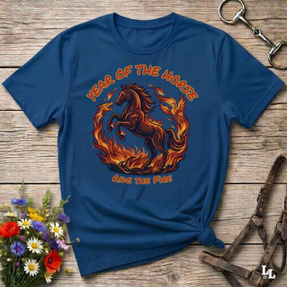 A navy blue graphic t-shirt featuring a fiery horse design with the text 'YEAR OF THE HORSE' and 'RIDE THE FIRE', laid flat on a rustic wooden background with decorative flowers and leather tack.