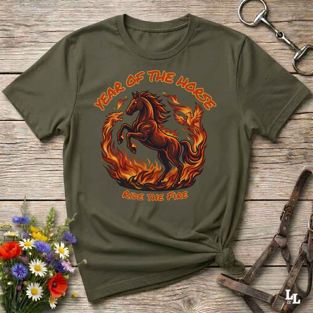 An olive green graphic t-shirt laid flat on a rustic wooden surface, styled with a horse halter, flowers, and a small knot at the bottom right. The t-shirt features a graphic of a fiery red and orange horse surrounded by flames, with the text 'YEAR OF THE HORSE' and 'RIDE THE FIRE'.