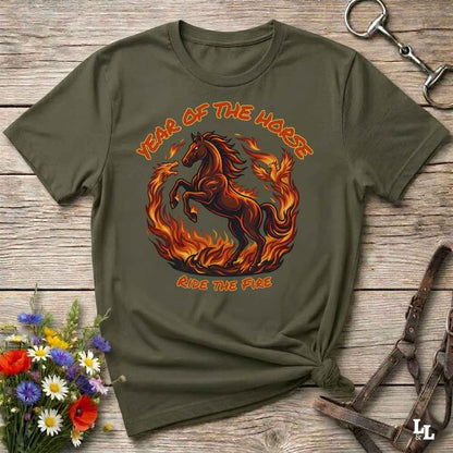 An olive green graphic t-shirt laid flat on a rustic wooden surface, styled with a horse halter, flowers, and a small knot at the bottom right. The t-shirt features a graphic of a fiery red and orange horse surrounded by flames, with the text 'YEAR OF THE HORSE' and 'RIDE THE FIRE'.