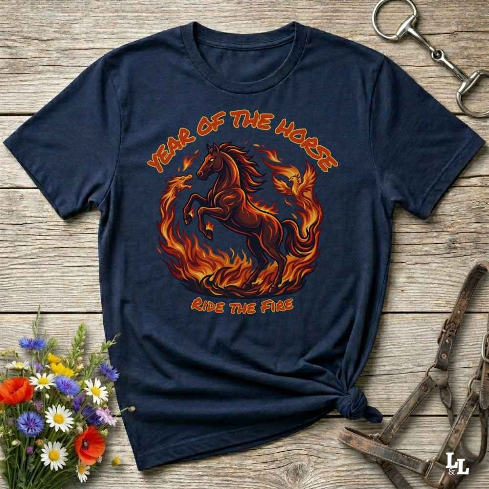 A navy blue graphic t-shirt featuring a fiery red and orange horse design with the text 'YEAR OF THE HORSE' and 'RIDE THE FIRE', laid flat on a rustic wooden background with decorative equestrian and floral props.