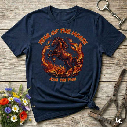 A navy blue graphic t-shirt featuring a fiery red and orange horse design with the text 'YEAR OF THE HORSE' and 'RIDE THE FIRE', laid flat on a rustic wooden background with decorative equestrian and floral props.