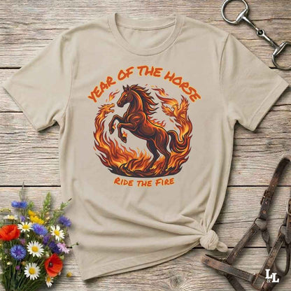 A beige graphic t-shirt featuring a fiery horse design with the text 'YEAR OF THE HORSE' and 'RIDE THE FIRE', styled on a rustic wooden background with flowers and leather tack.