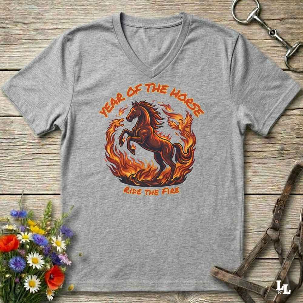 grey V-neck t-shirt laid flat on a rustic wooden surface, featuring a graphic of a rearing horse surrounded by flames with the text 'YEAR OF THE HORSE' and 'RIDE THE FIRE'. The shirt is styled with equestrian props including a leather bridle, horse bits, and a small bouquet of wildflowers.