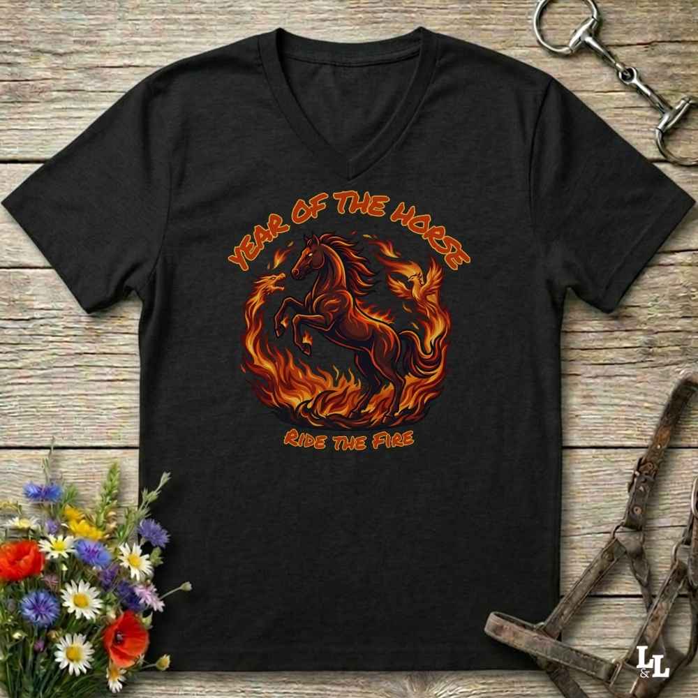 black V-neck t-shirt laid flat on a rustic wooden surface, featuring a graphic of a rearing horse surrounded by flames with the text 'YEAR OF THE HORSE' and 'RIDE THE FIRE'. The shirt is styled with equestrian props including a leather bridle, horse bits, and a small bouquet of wildflowers.